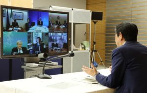 A speaker siting in front  of the camera, on a video conference. In fron t of him a Zoom screen with 6 other participants on a video conference call.